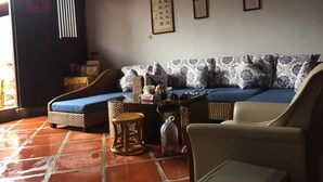 Ein Wohnzimmer mit einem blauen Sofa, einem Korbstuhl, einem kleinen runden Tisch und einem Fliesenboden.