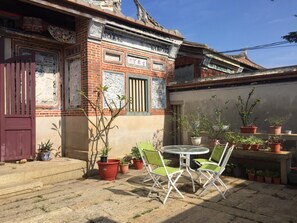 Une cour avec un bâtiment en briques, une petite table ronde entourée de chaises et diverses plantes en pot.