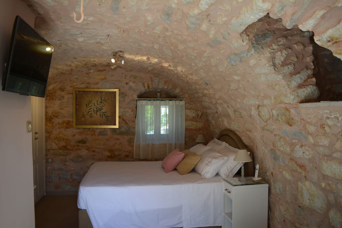 Ein Schlafzimmer mit Steinkamin, einem Bett mit weißer Bettwäsche, einem Nachttisch mit Lampe und einem an der Wand montierten Fernseher.