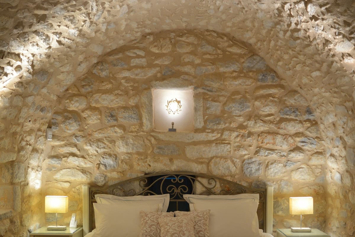 Ein Schlafzimmer mit steinverzieltem Gewölbedecke, einem großen Bett mit vielen Kissen, Nachttischlampen und einer hölzernen Bank.
