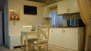 A compact kitchen with white cabinets, a dining table with two chairs, a television mounted above the counter, and a stone wall. - Olea (East Mani)