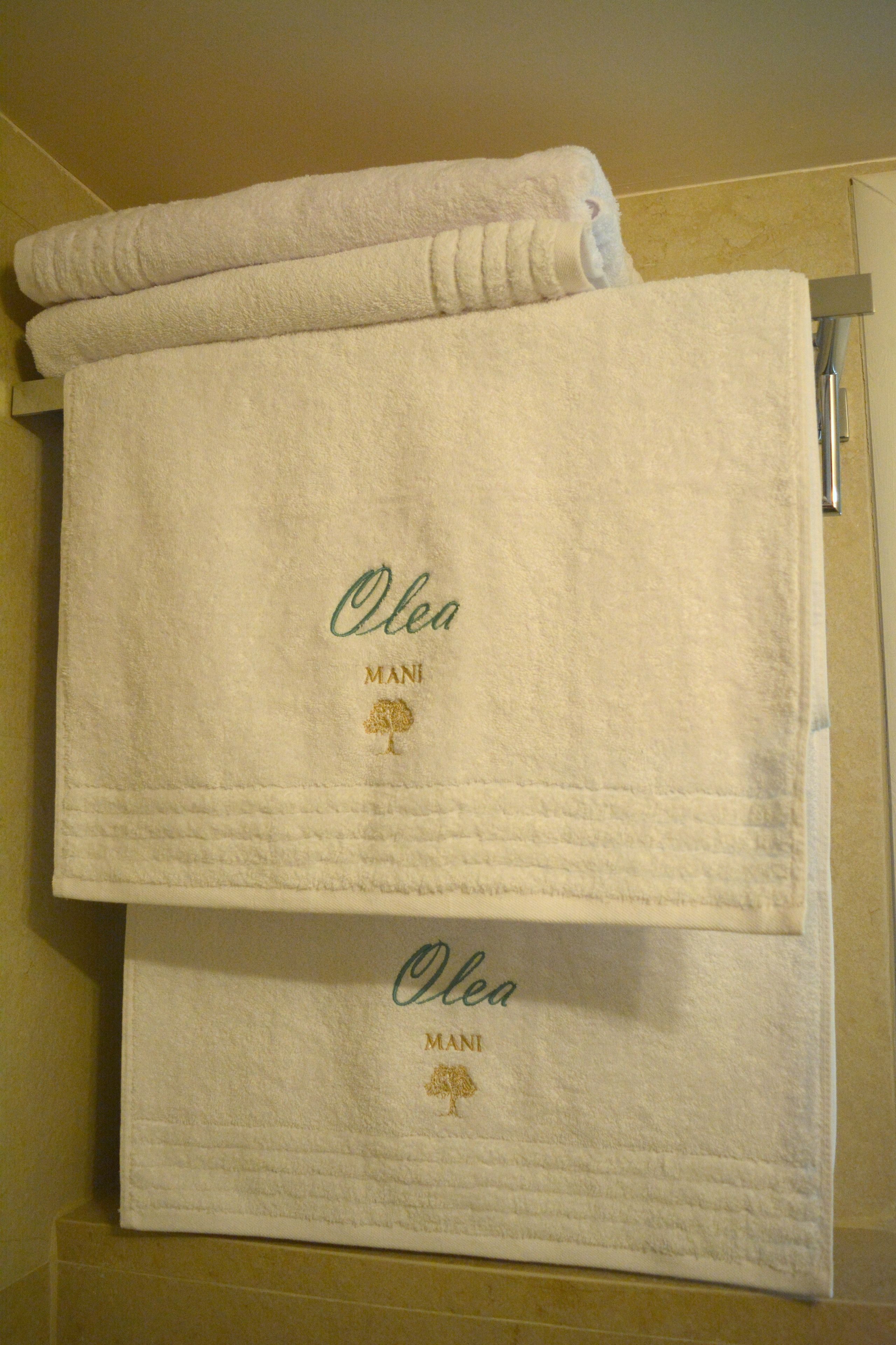Ein Satz weißer Handtücher mit eingesticktem Text und einem Baum-Logo, die an einem Haken im Badezimmer hängen.