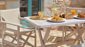 An outdoor dining area with stone walls, a table with plates of food, and a chair. - Olea (East Mani)