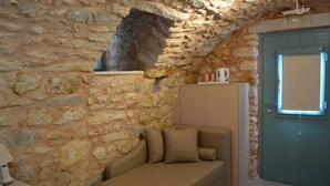 A stone-walled room with a beige sofa, a small table with a lamp, and a door with a window. - Olea (East Mani)