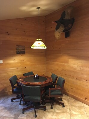 Ein Zimmer mit Holzwänden, einem runden Tisch und sechs Stühlen. Eine Hängeleuchte ist über dem Tisch zu sehen.
