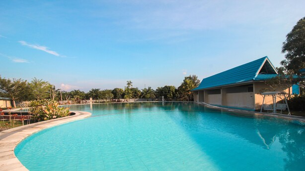 A clear blue swimming pool with a curved edge, surrounded by a tiled deck and a building with a blue roof. - OYO 1153 Tiga Dara Hotel & Resort Syariah (Pekanbaru)