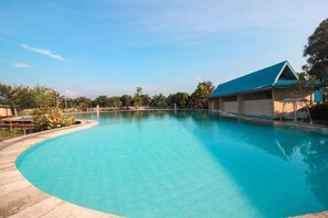 A clear blue swimming pool with a curved edge, surrounded by a tiled deck and a building with a blue roof. - OYO 1153 Tiga Dara Hotel & Resort Syariah (Pekanbaru)