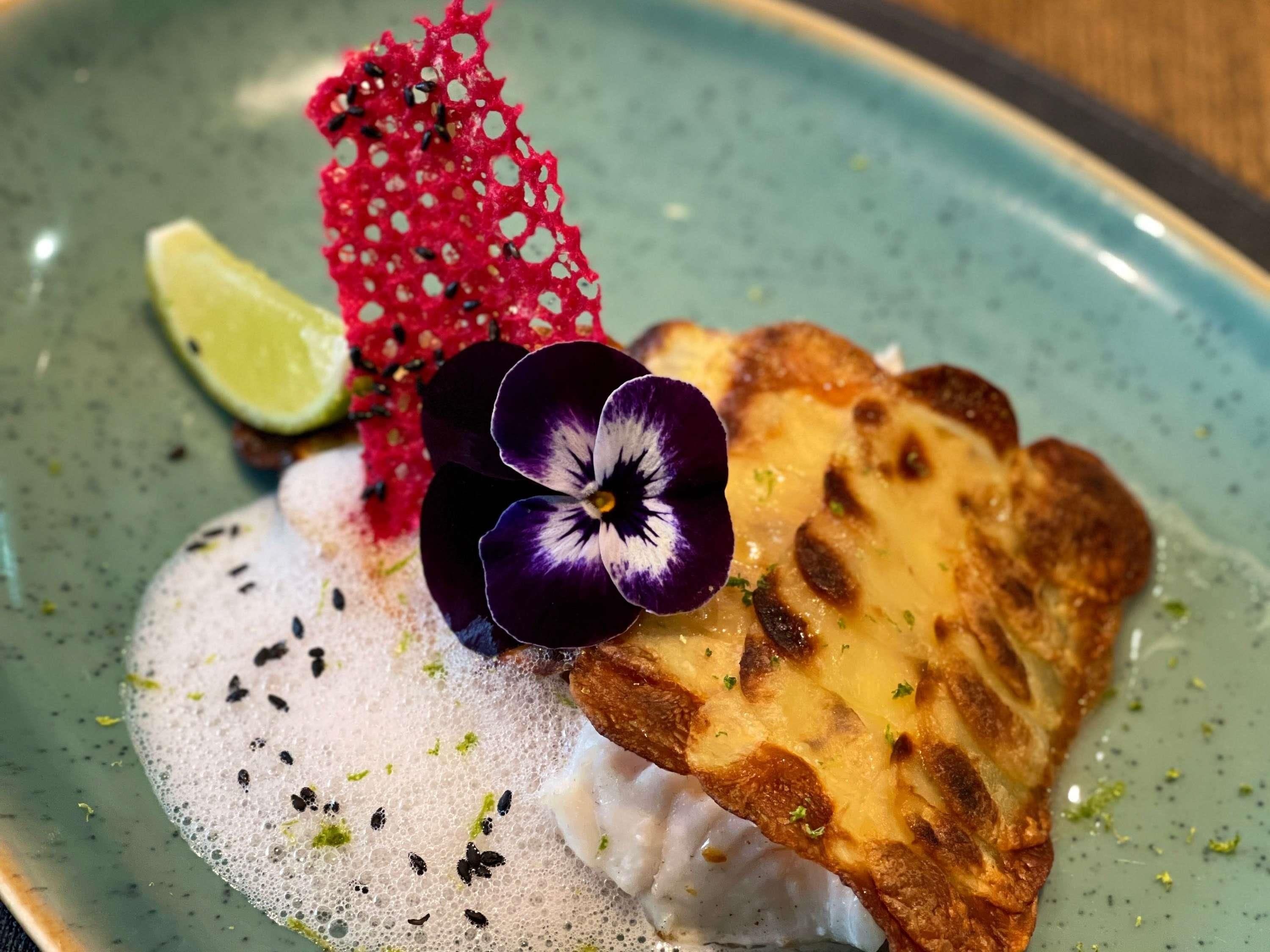 A plate with a piece of grilled fish, a slice of red fruit, a purple flower, and a lime wedge.