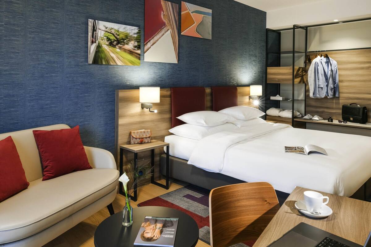 A hotel room with a bed, a sofa, a desk with a laptop, a chair, a small table with a cup, and a nightstand with a lamp.