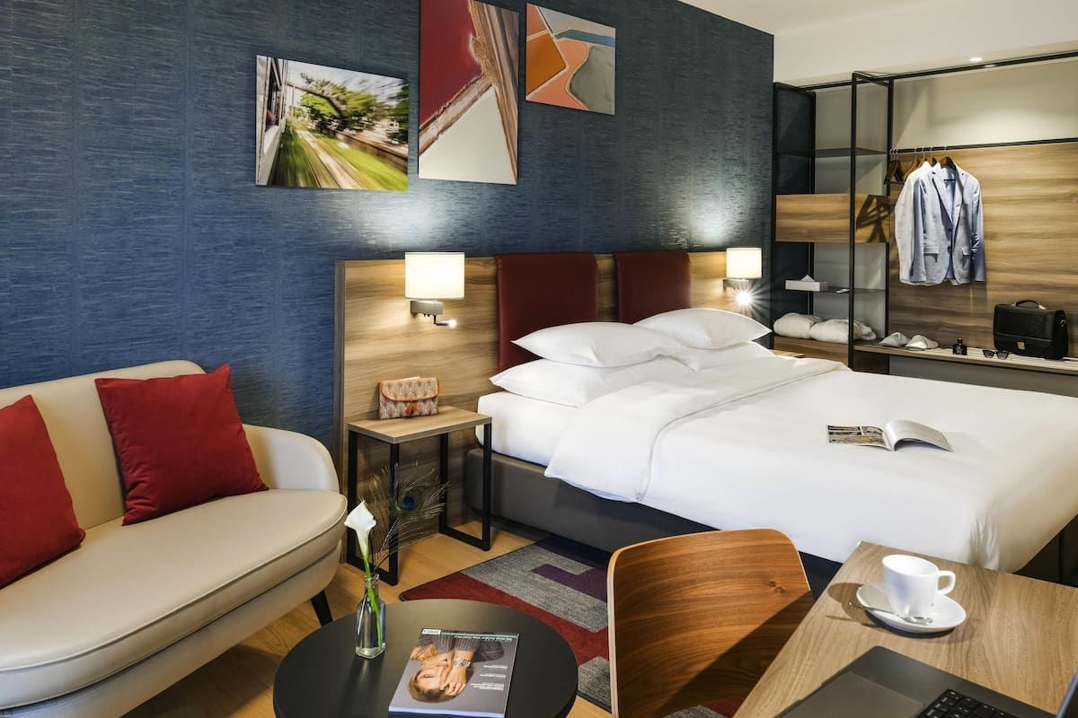 A hotel room with a bed, a sofa, a desk with a laptop, a chair, a small table with a cup, and a nightstand with a lamp.