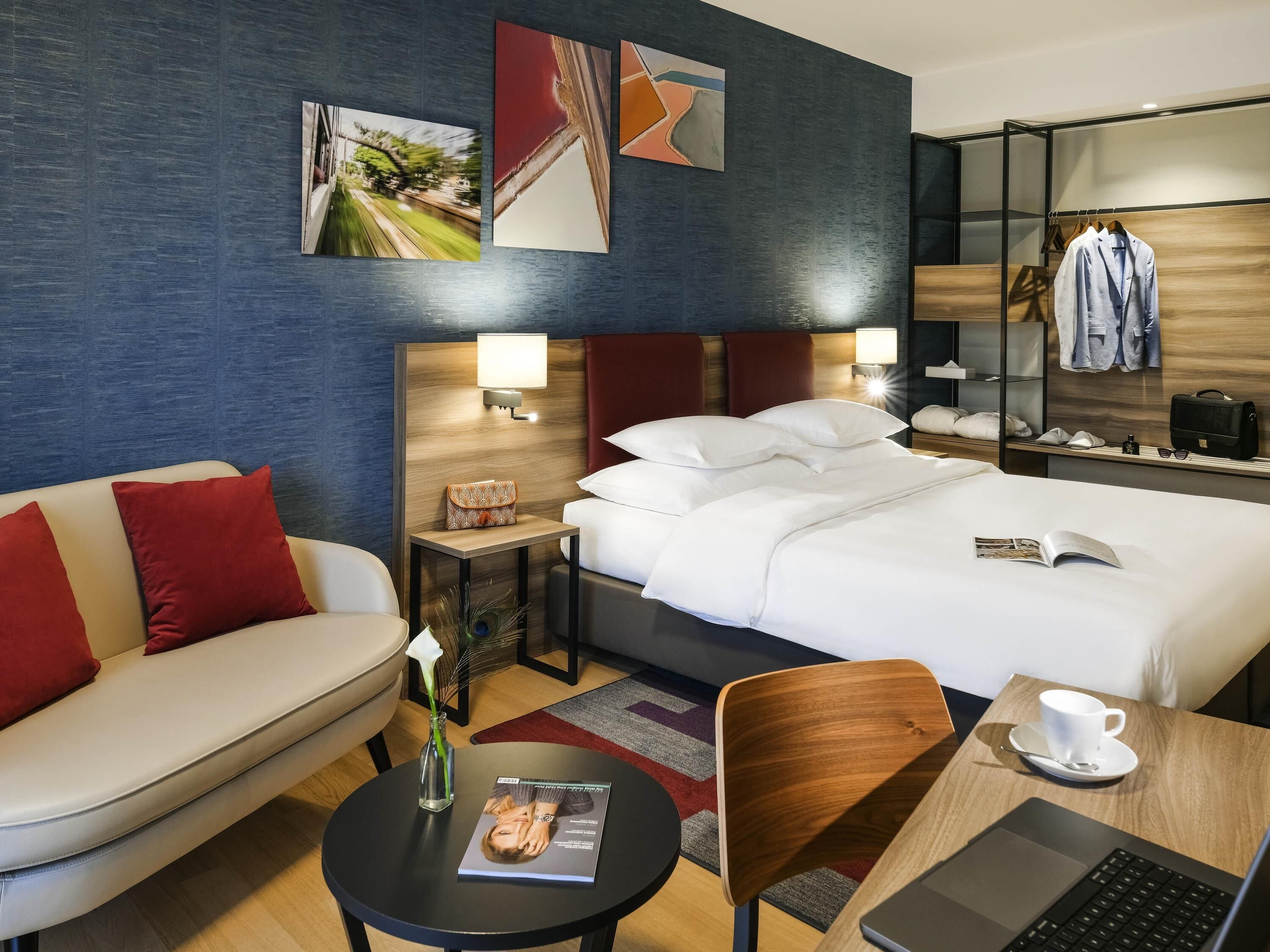 A hotel room with a bed, a sofa, a desk with a laptop, a chair, a small table with a cup, and a nightstand with a lamp.