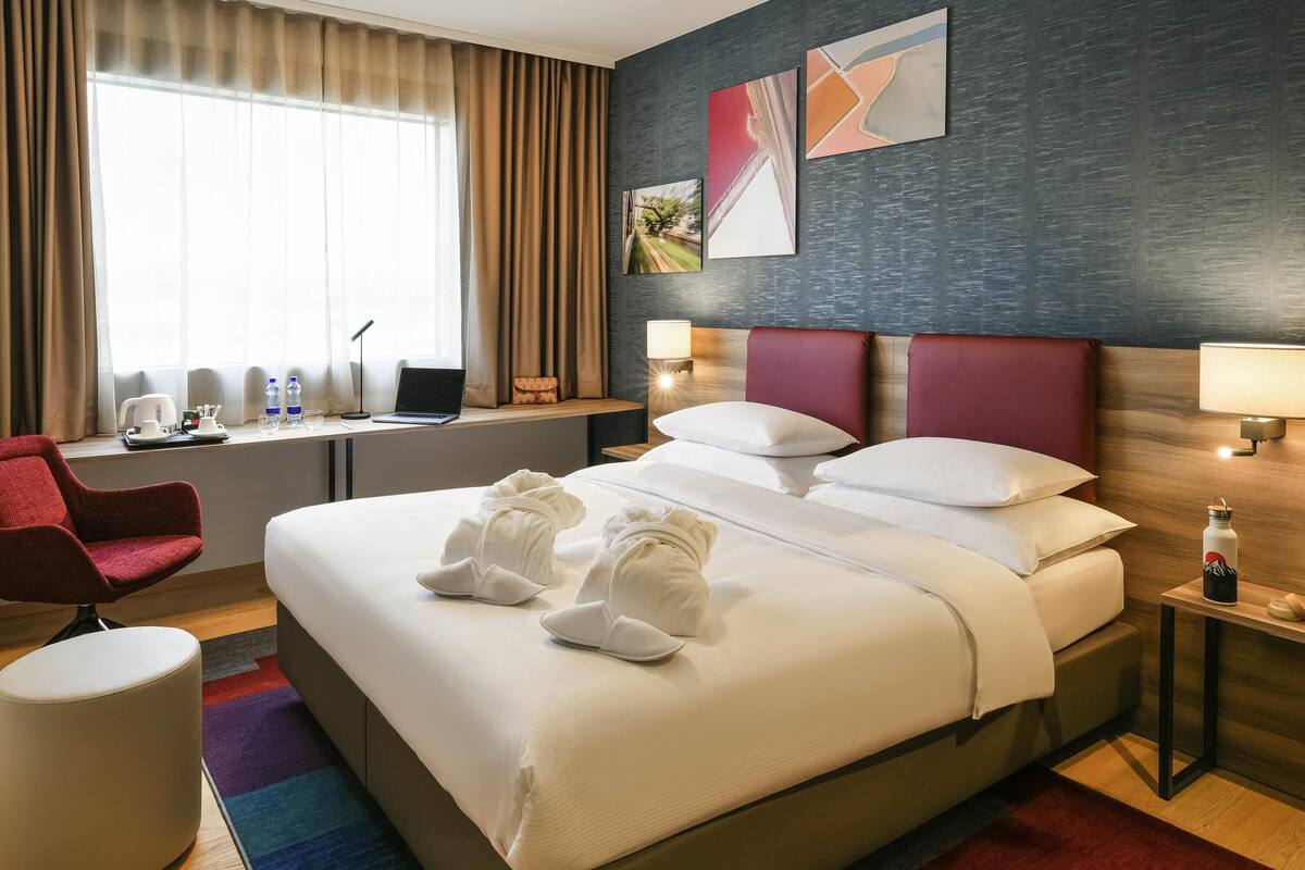 A hotel room with a large bed, a desk with a laptop, a red chair, a round ottoman, and a window with curtains.