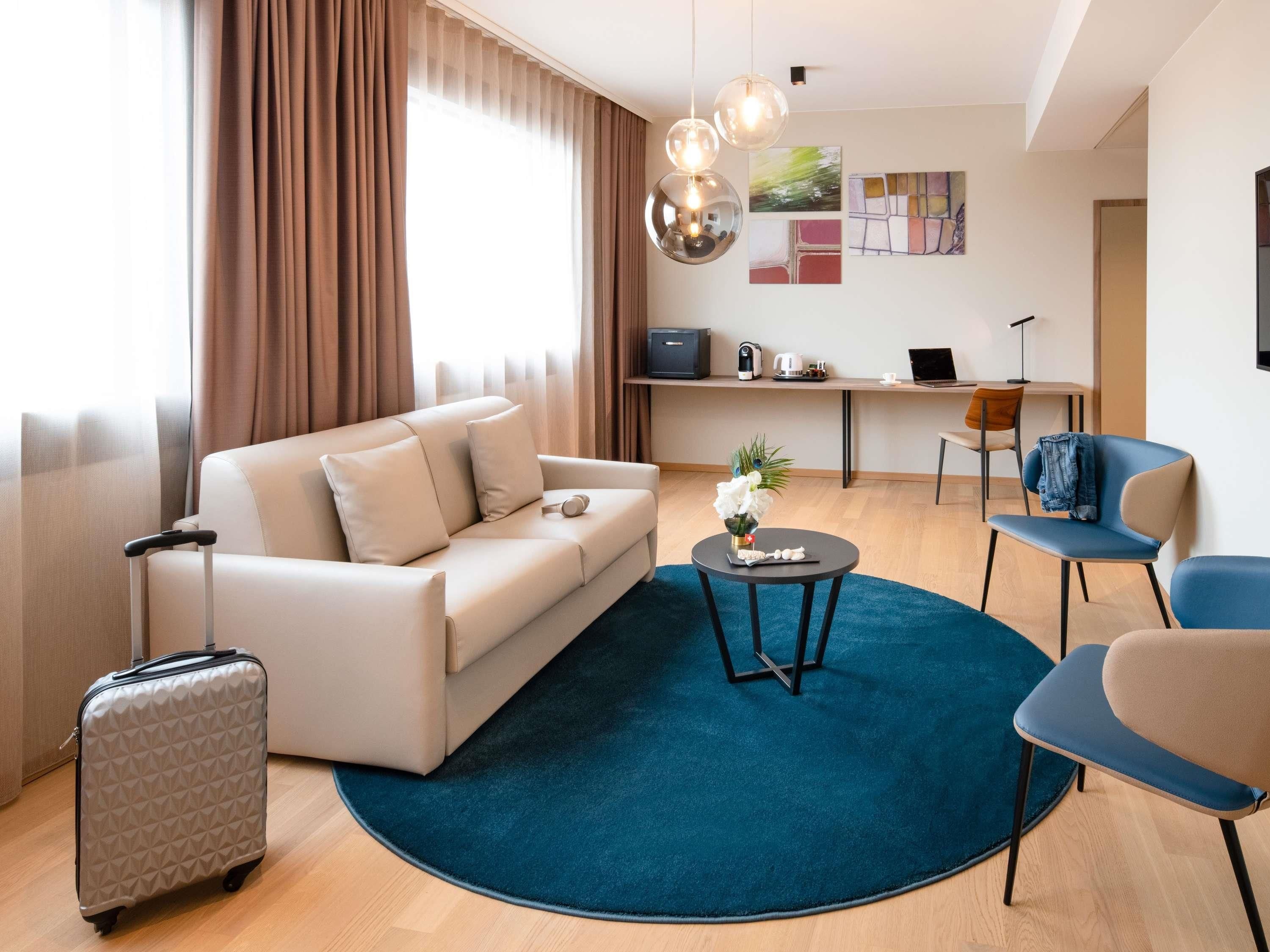 A modern hotel room with a sofa, a small round table, a desk, and a chair. There is a suitcase and a plant on the floor.