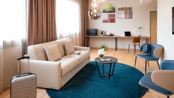 A modern hotel room with a sofa, a small round table, a desk, and a chair. There is a suitcase and a plant on the floor.