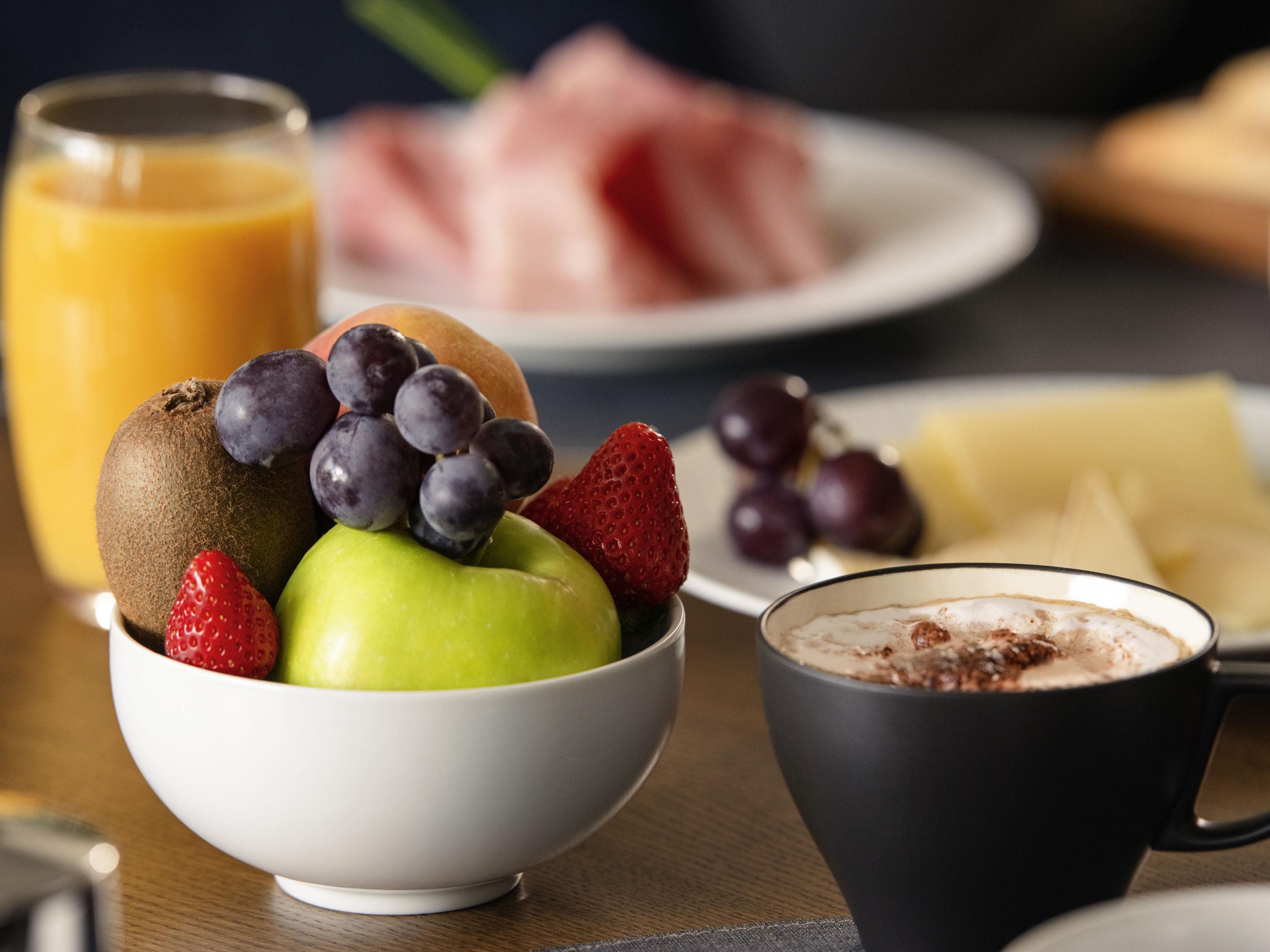 Un desayuno con un tazón de fruta, un vaso de jugo de naranja, un plato de jamón, una taza de café y una rebanada de queso.