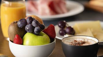 Un desayuno con un tazón de fruta, un vaso de jugo de naranja, un plato de jamón, una taza de café y una rebanada de queso.