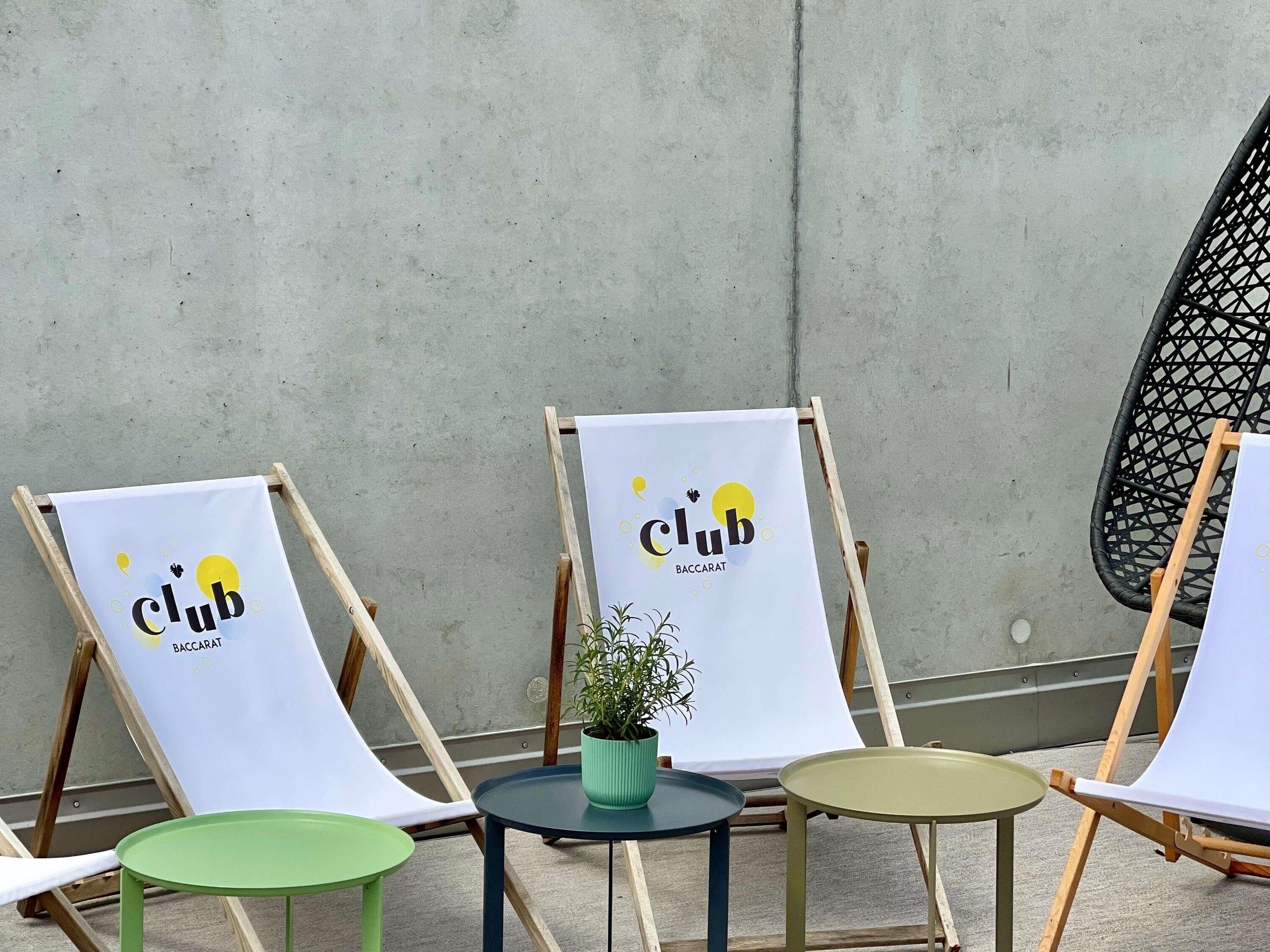 Tres sillas de lona con el logotipo 'Club Baccarat', una pequeña mesa redonda con una planta en maceta y una silla tejida de color negro frente a una pared de hormigón.