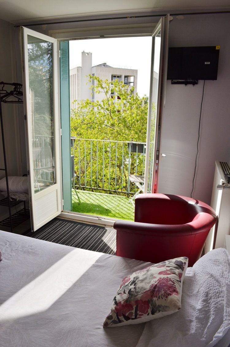Una habitación con una cama, una silla roja y un balcón con vistas a edificios y árboles.