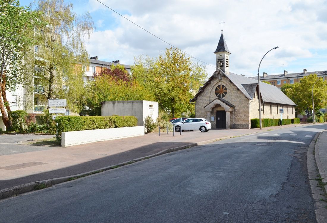 Una iglesia con campanario y reloj, un coche blanco estacionado al frente, y un edificio residencial al fondo.