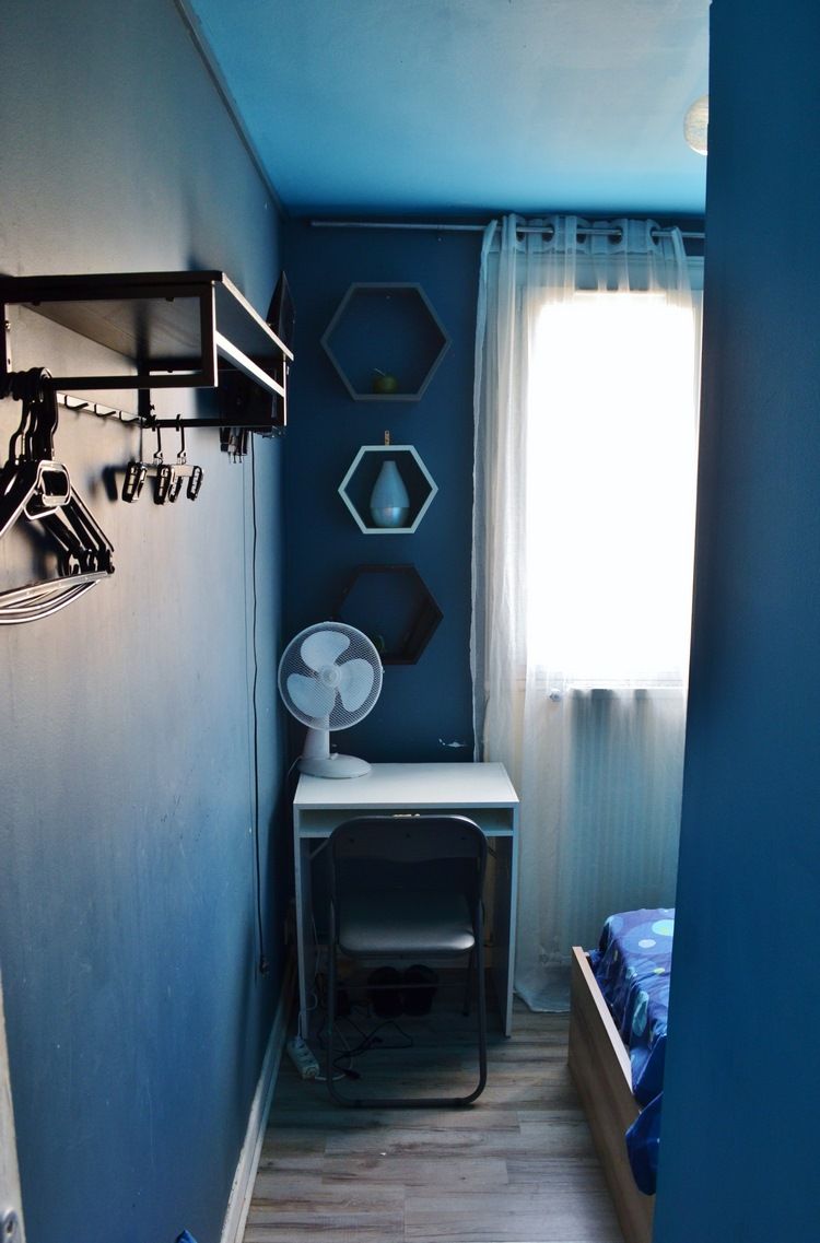 Quarto pequeno e estreito com uma cama, uma escrivaninha e uma cadeira. Há uma janela com cortinas, um ventilador e prateleiras fixadas na parede com diversos objetos.