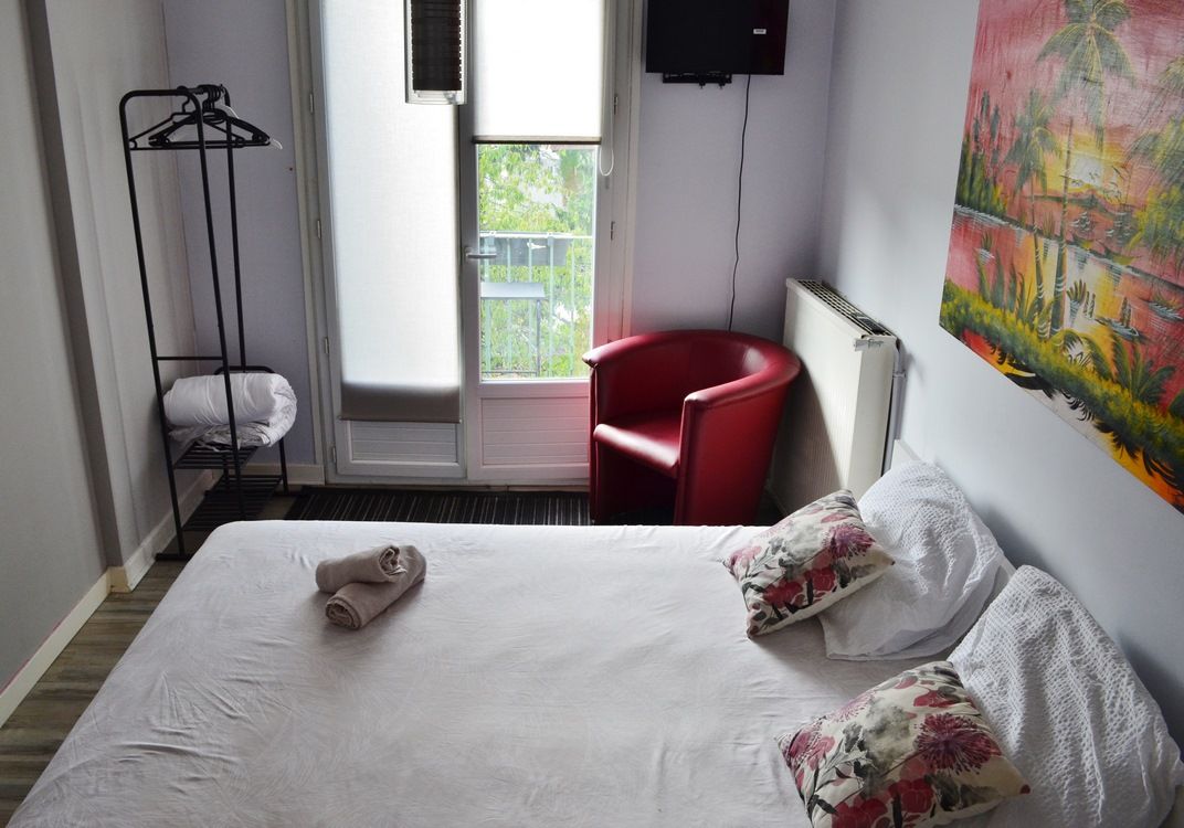 Hotellihuone, jossa on sänky, punainen tuoli, naulakko ja taulu seinällä.