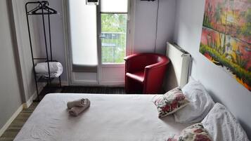 Hotellihuone, jossa on sänky, punainen tuoli, naulakko ja taulu seinällä.