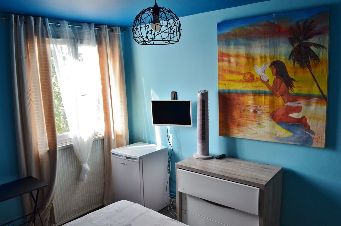 A room with a bed, a small white cabinet, a TV, a standing fan, and a painting on the wall.