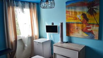 A room with a bed, a small white cabinet, a TV, a standing fan, and a painting on the wall.