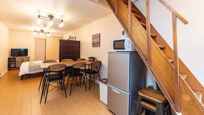 Un espacio habitable compacto con una escalera de madera, un comedor con mesa y sillas, un refrigerador y un televisor al fondo.
