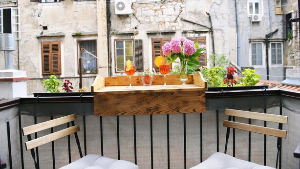 A balcony with a wooden table holding drinks and flowers, overlooking a historic building with stone walls and wooden shutters. - Vitopolis Apartment 3 (Rijeka)