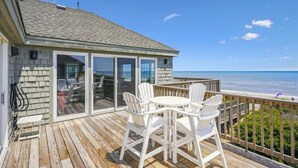 A wooden deck with a table and chairs, a glass door leading to a room, and a view of the ocean. - Oceanfront at Ships Watch! (Duck)