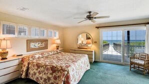 A bedroom with a bed, bedside tables, a chair, a ceiling fan, and a view of the ocean through a sliding glass door. - Oceanfront at Ships Watch! (Duck)