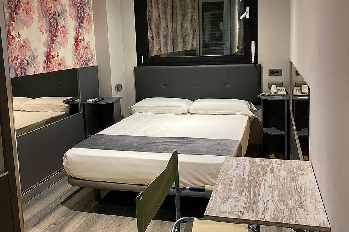 A modern hotel room with a large bed, a bedside table with a lamp, a small desk with a chair, and a large mirror above the bed.