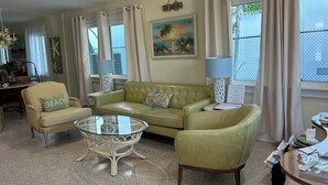 Un salon avec un canapé vert, une table basse en verre et un fauteuil orné d’un coussin sur lequel est écrit le mot « PLAGE ». On y trouve deux lampes, un tableau et une fenêtre avec des rideaux.