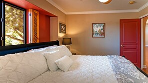 A bedroom with a large bed, a window with a view of trees, a door, and a painting on the wall.