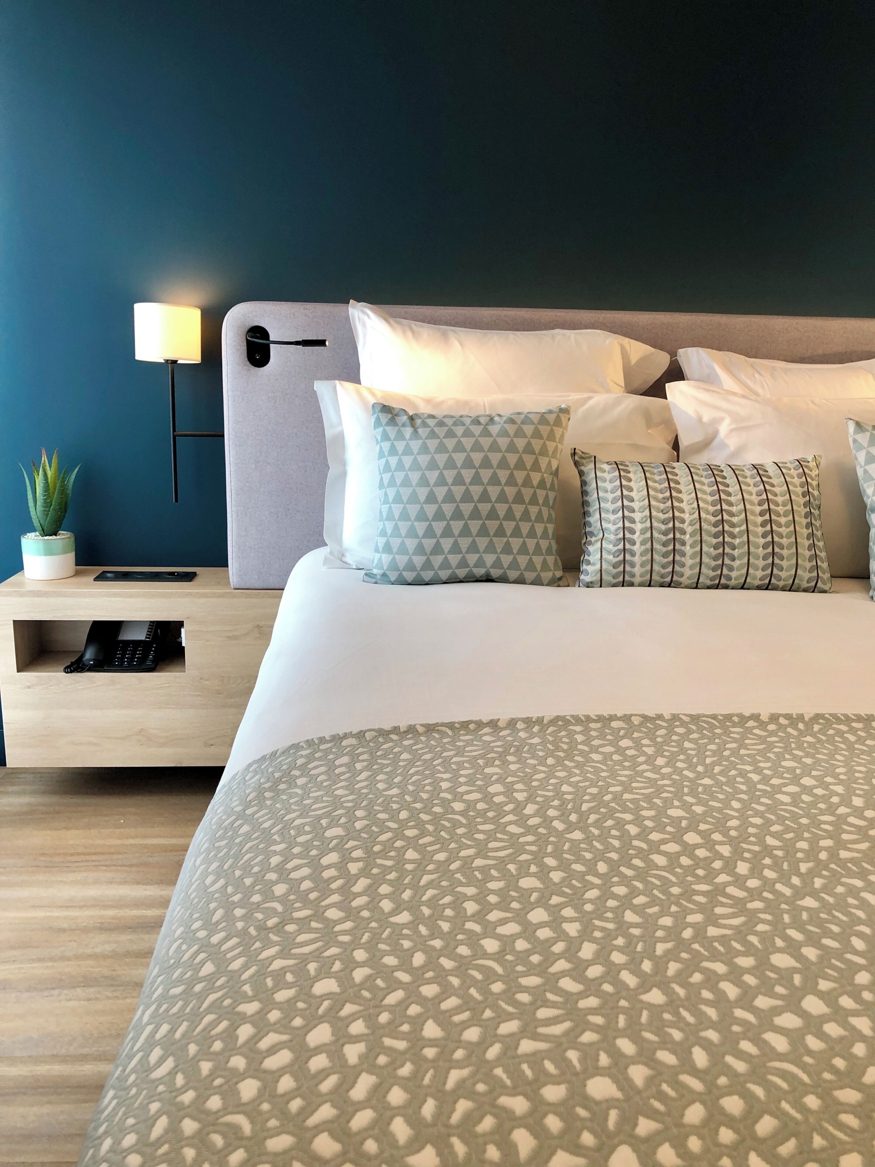 A neatly made bed with patterned bedding, a bedside table with a lamp, a small plant, and a keyboard.