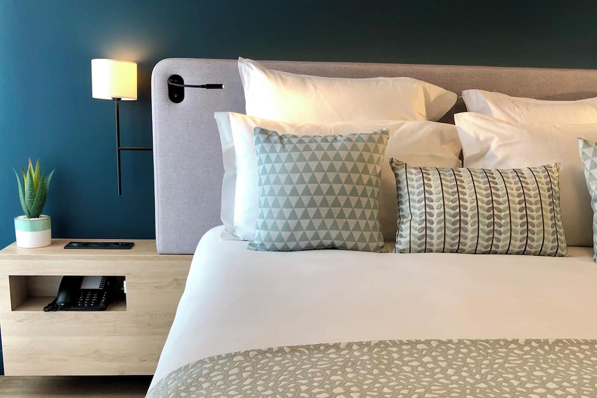 A neatly made bed with patterned bedding, a bedside table with a lamp, a small plant, and a keyboard.
