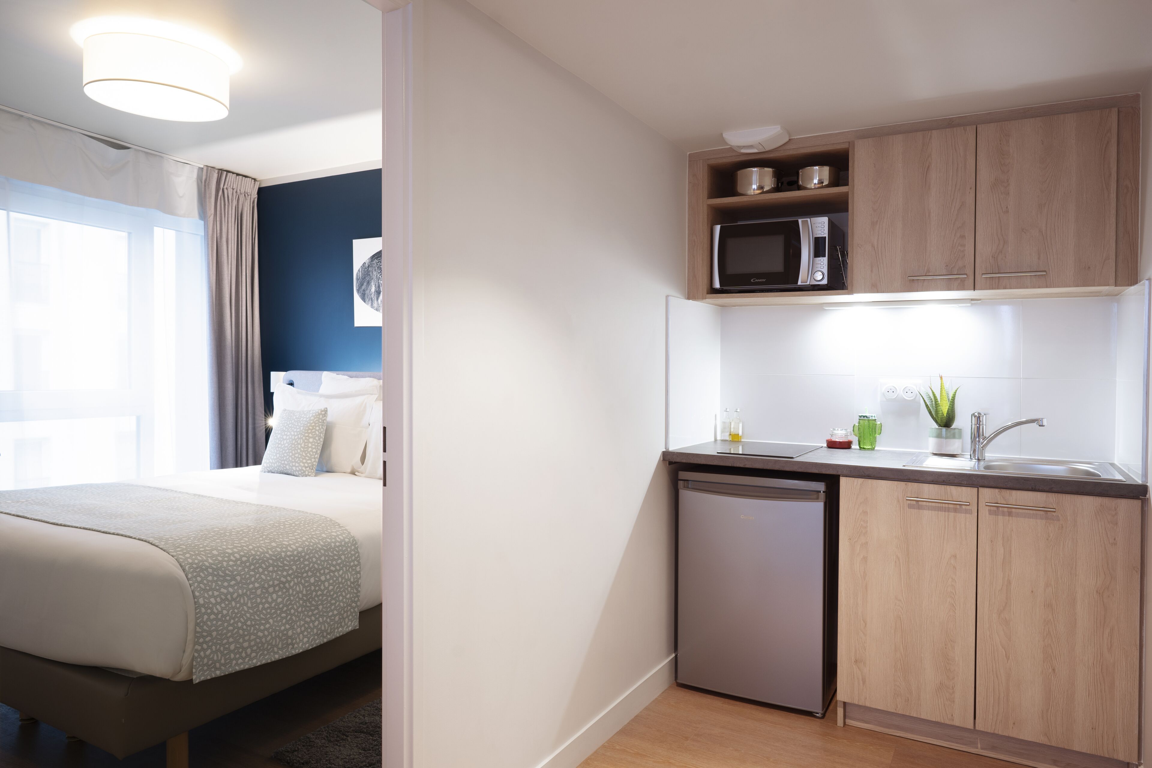 A modern hotel room with a bed, a small kitchenette, and a view of the outdoors.