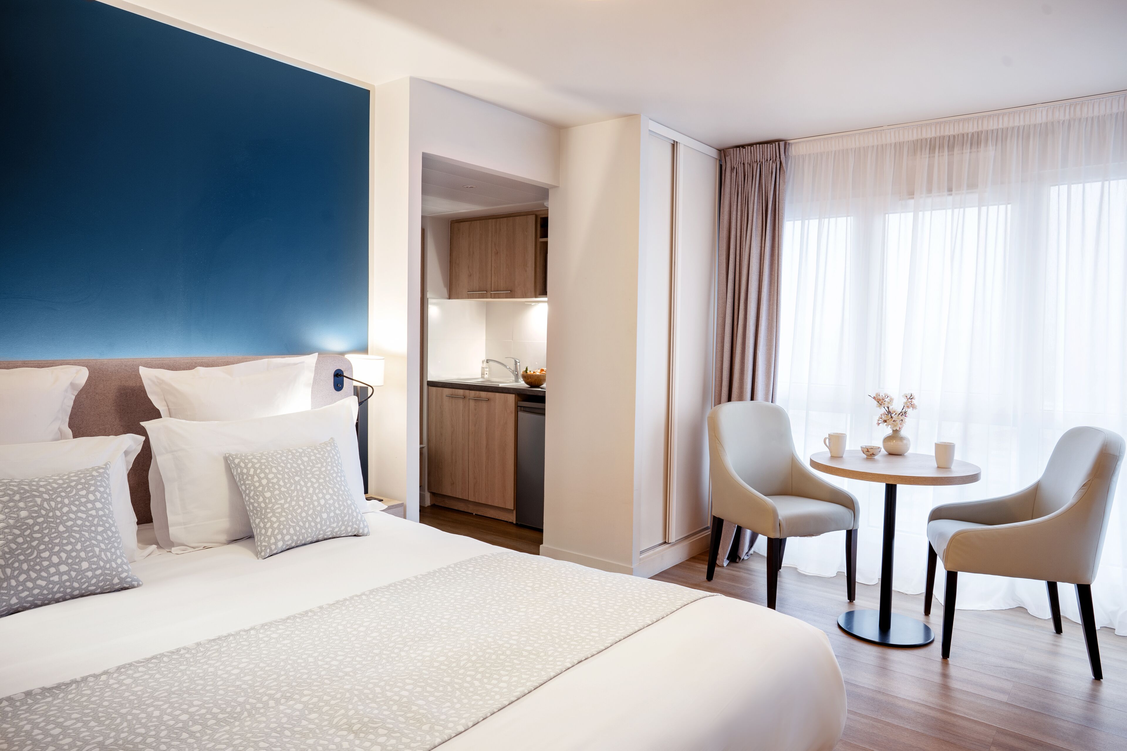 Een hotelkamer met een groot bed, een kleine kitchenette, twee stoelen rond een tafel en uitzicht op de omgeving door dunne gordijnen.
