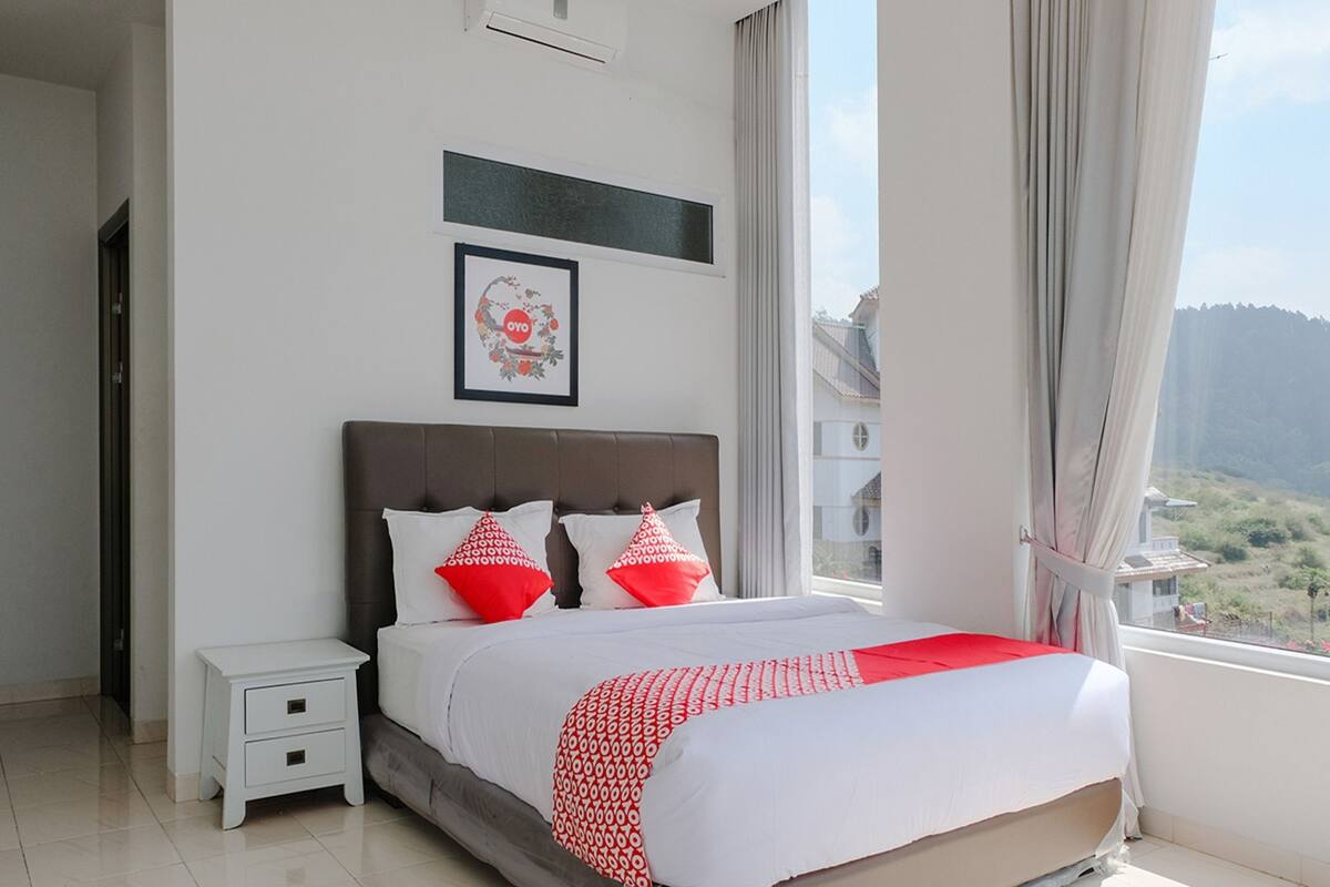 Une chambre à coucher avec un grand lit, une table de chevet, un tableau encadré au mur et une fenêtre avec des rideaux.
