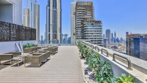 A rooftop terrace with seating, a view of modern high-rise buildings, and a landscaped area with plants. - Driven Holiday Homes - Mada Residences (Dubai)