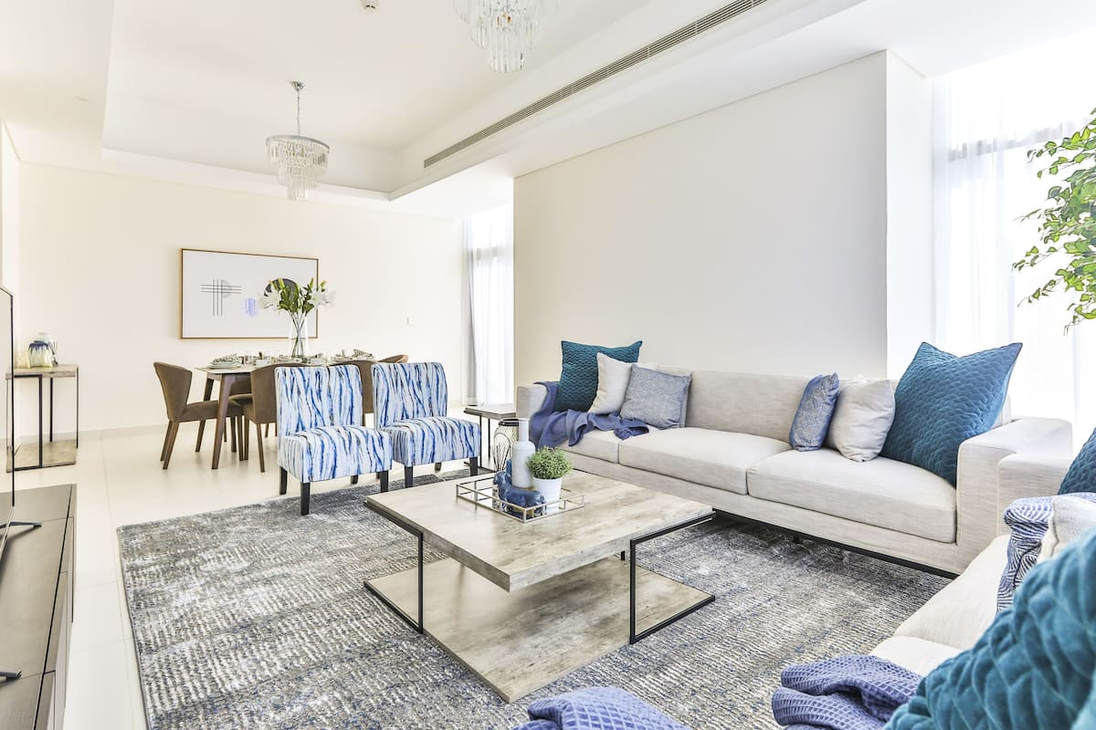 A modern living room with a sofa, coffee table, and chairs. There is a dining area with a table and chairs in the background.