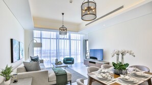 A modern living room with a sofa, dining table set for four, and a large window with curtains. - Driven Holiday Homes - Mada Residences (Dubai)