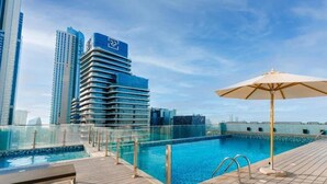 A rooftop pool with a glass barrier, a wooden deck, and an umbrella. - Driven Holiday Homes - Mada Residences (Dubai)