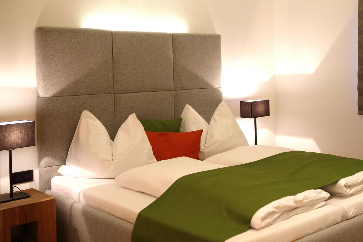 Una cama bien hecha con cabecera gris, ropa de cama blanca y verde, y una lámpara de noche.