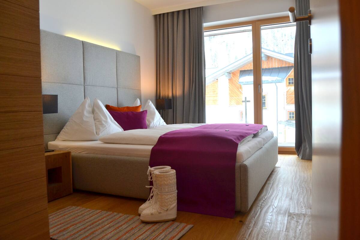 Una habitación de hotel moderna con una cama, una mesita de noche, un ventanal con cortinas y vistas a un edificio.