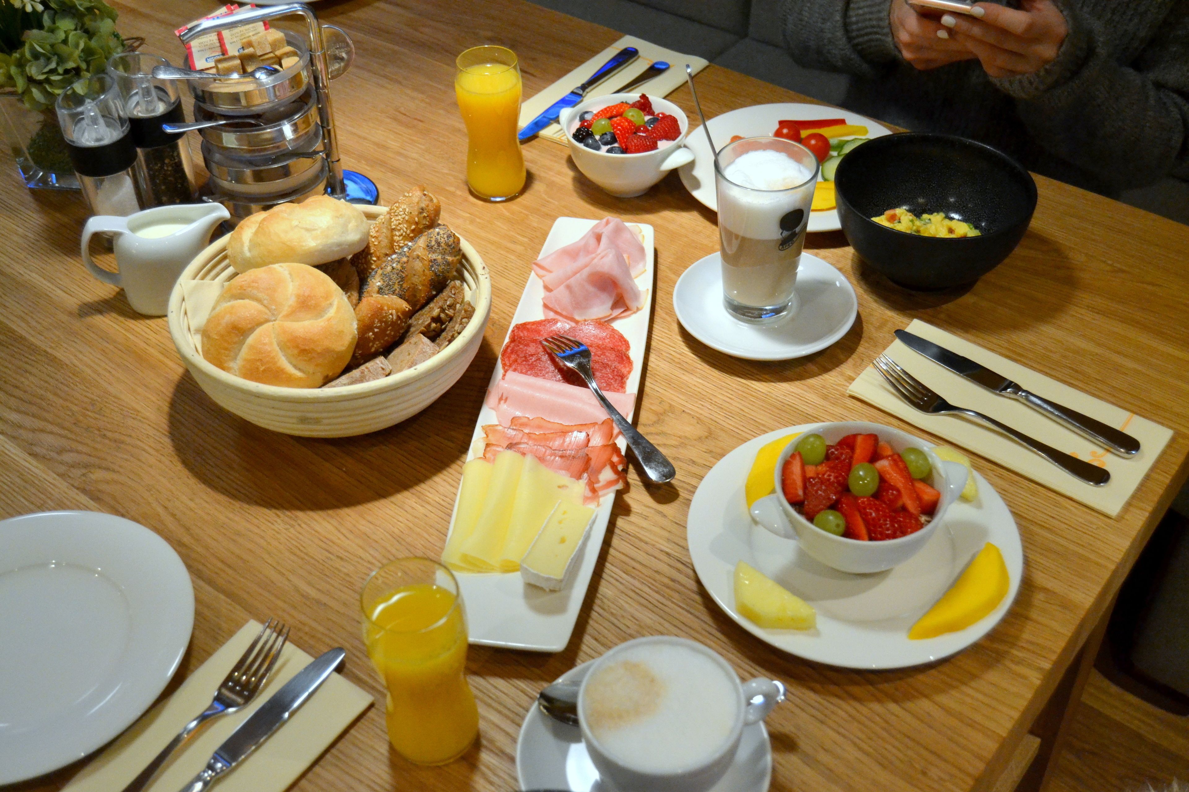 Una mesa de desayuno con diversos platos, incluyendo pan, jamón, queso y fruta.