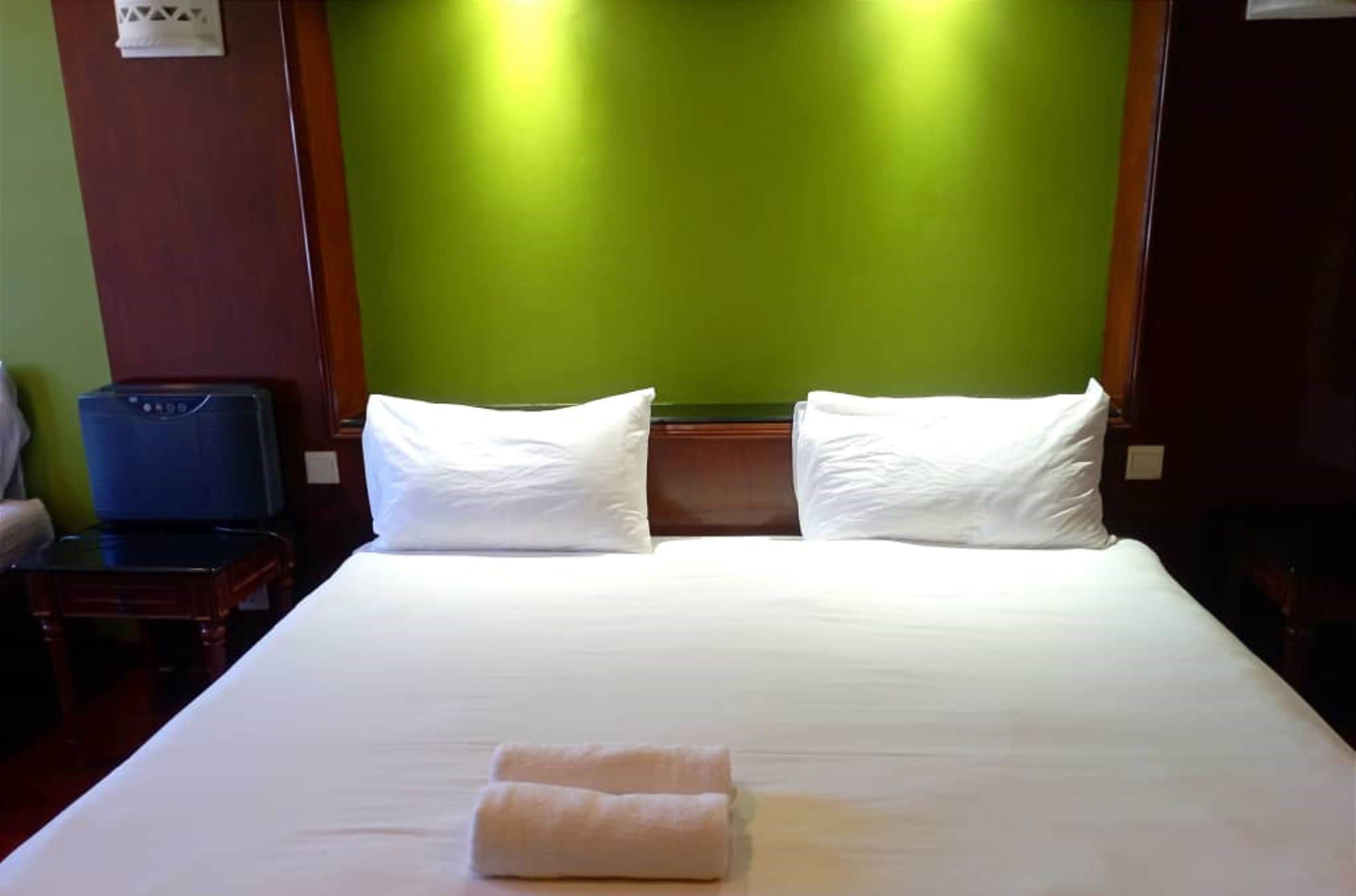 Habitación de hotel con una cama grande, dos almohadas, una toalla doblada, un cabecero de madera y una maleta azul sobre una mesita de noche de madera.