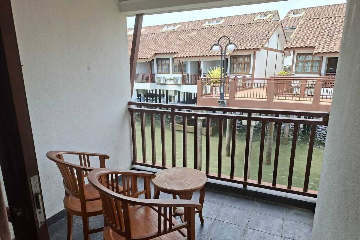 Varanda com duas cadeiras de madeira e uma mesa pequena, com vista para um jardim e edifícios com telhados de azulejo.
