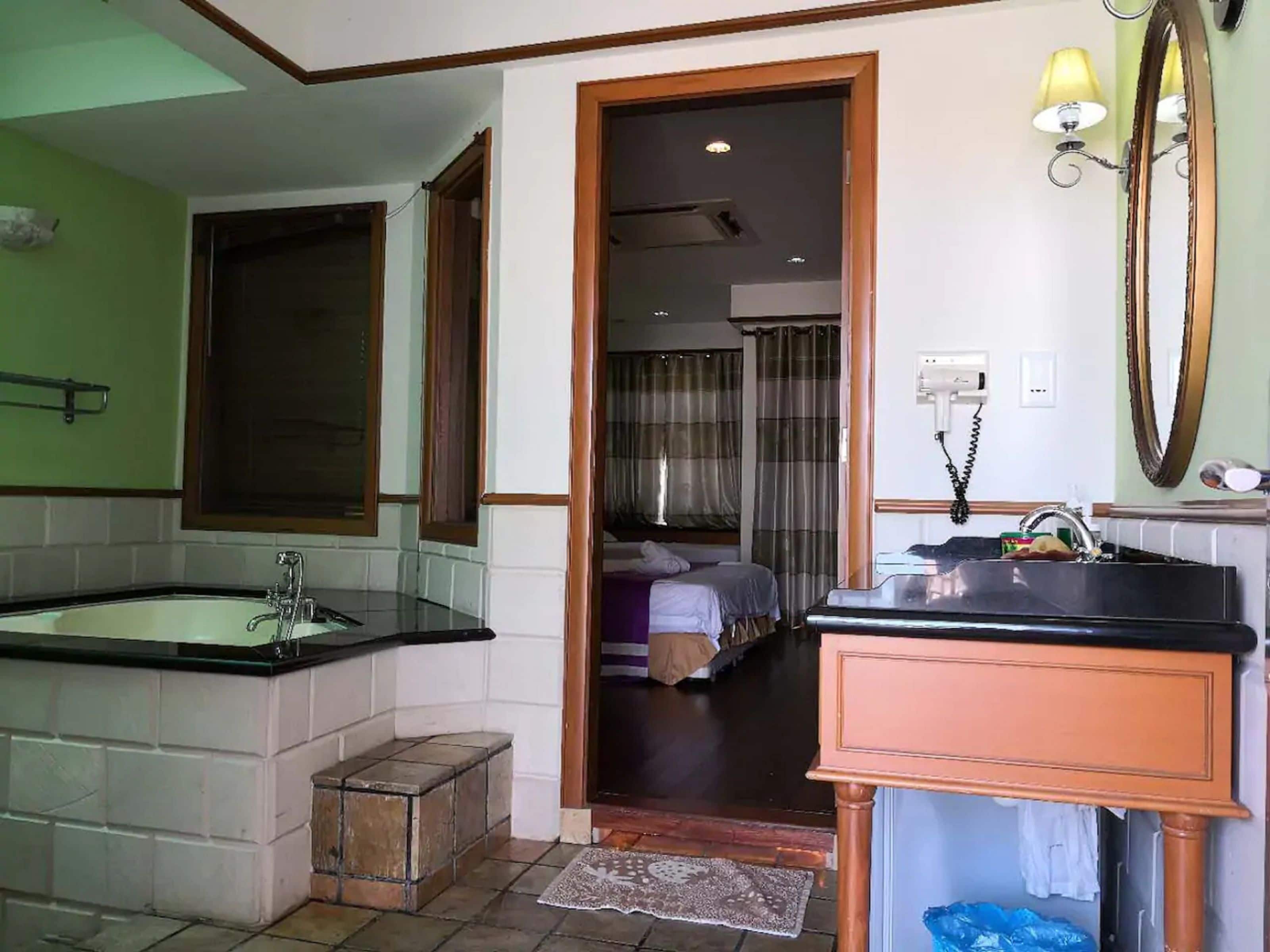 A bathroom with a large bathtub, a sink, and a mirror. There is a door leading to a bedroom with a bed and curtains.